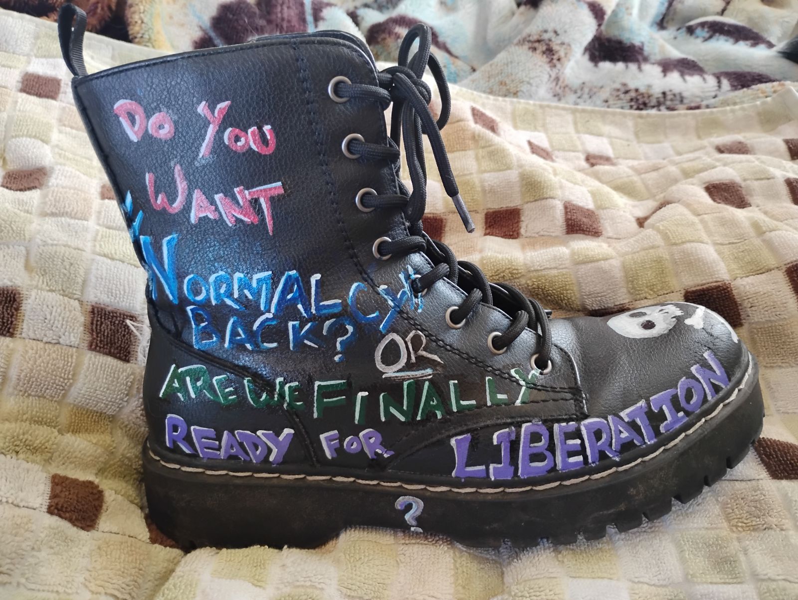 A black leather boot with graffiti-style writing in purple, pink, and blue that reads: 'do you want normalcy or are we finally ready for liberation'