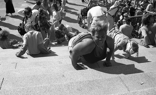 A black and white photograph of a femme-presenting disability rights protester with disheveled hair crawling up the stone steps of the U.S. Capitol during the 1990 Capitol Crawl, visibly exhausted from the physical exertion of the protest
